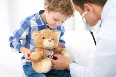 A little boy is holding a stuffed bear and the doctor is helping the little boy to feel comfortable by using a stethoscope to listen for the stuffed bears heart
